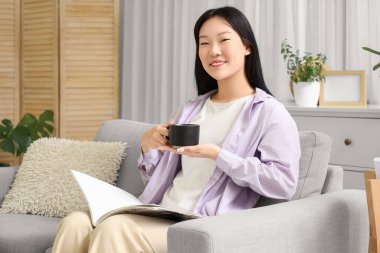 Genç Asyalı kadın oturma odasında kahve içiyor ve dergi okuyor.