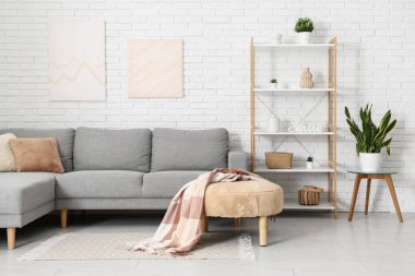 Beyaz tuğla duvarın yanında gri kanepesi ve raf ünitesi olan aydınlık oturma odasının içinde.