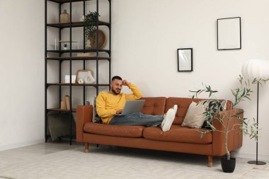 Modern dizüstü bilgisayarlı genç adam oturma odasında kahverengi deri koltukta oturuyor.