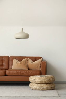Beyaz duvarın yanında minderleri ve pufları olan kahverengi deri kanepe.