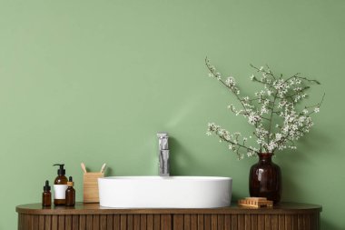 Çiçek açan dalları olan vazo, banyo malzemeleri ve banyodaki yeşil duvarın yanındaki çekmecelere batan lavabo.