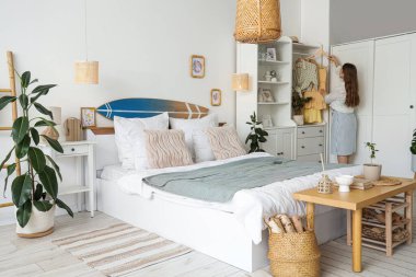 Yatak odasında dolaptan giysilerini ve sörf tahtasını alan genç bir kadın.