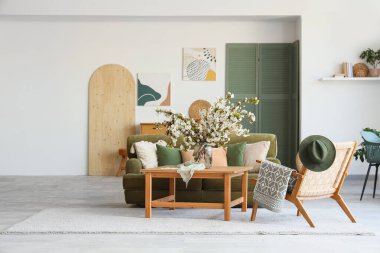 Yeşil kanepesi, koltuğu ve vazoda çiçekli dalları olan aydınlık oturma odasının içinde.