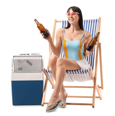 Portatif buzdolabı ve soğuk birayla beyaz arka planda plaj sandalyesinde oturan genç bir kadın.