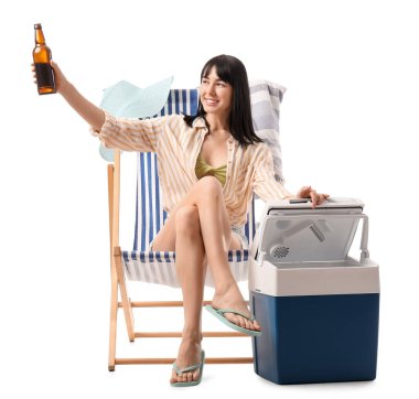 Portatif buzdolabı ve soğuk birayla beyaz arka planda plaj sandalyesinde oturan genç bir kadın.