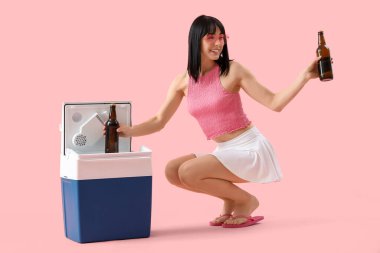 Portatif buzdolabı ve pembe arka planda soğuk bira şişeleri olan genç bir kadın.