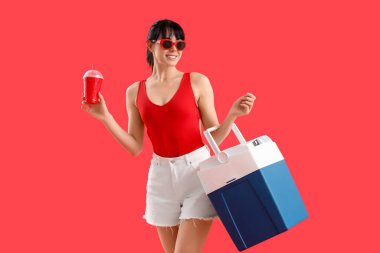 Portatif buzdolabı ve kırmızı arka planda bir fincan soğuk içeceği olan genç bir kadın.