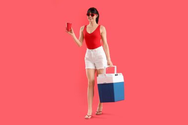 Portatif buzdolabı ve kırmızı arka planda bir fincan soğuk içeceği olan genç bir kadın.