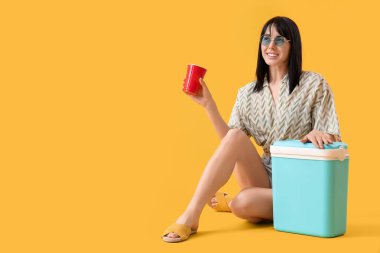 Portatif buzdolabı ve sarı arka planda bir fincan soğuk içeceği olan genç bir kadın.