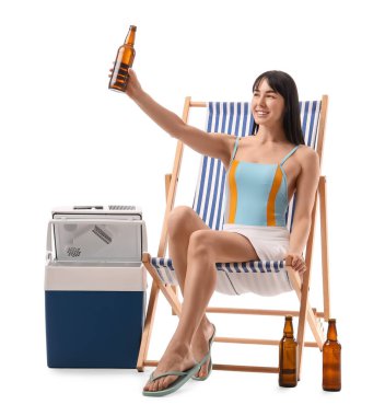 Portatif buzdolabı ve soğuk birayla beyaz arka planda plaj sandalyesinde oturan genç bir kadın.