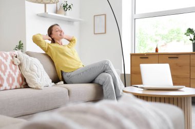 Oturma odasındaki gri kanepede dinlenen gülümseyen genç kadın.