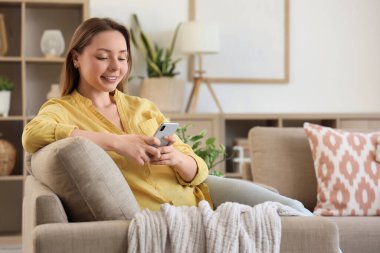 Oturma odasındaki gri koltukta cep telefonu kullanan gülümseyen genç kadın.