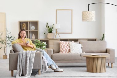 Oturma odasındaki gri koltukta cep telefonu kullanan gülümseyen genç kadın.