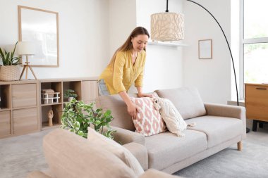 Genç bir kadın oturma odasındaki gri koltukta yastığı ayarlıyor.