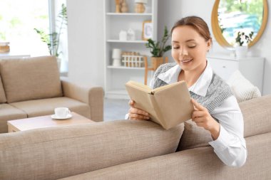 Gülümseyen genç kadın oturma odasındaki gri koltukta kitap okuyor.