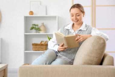 Oturma odasındaki gri koltukta kitap okuyan gülümseyen genç kadın.