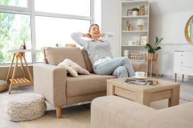 Oturma odasındaki gri kanepede dinlenen gülümseyen genç kadın.