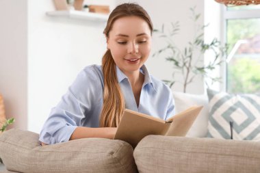 Oturma odasındaki gri koltukta kitap okuyan gülümseyen genç kadın.