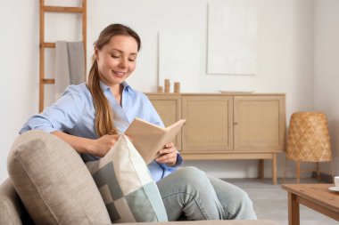 Oturma odasındaki gri koltukta kitap okuyan gülümseyen genç kadın.