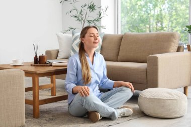 Sakin genç bayan oturma odasındaki gri kanepenin yanında meditasyon yapıyor.