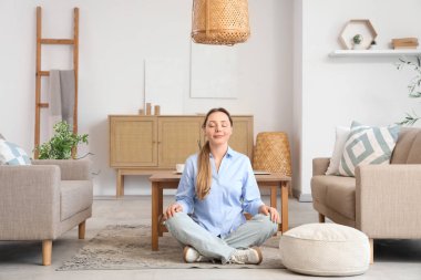 Sakin genç bayan oturma odasındaki gri kanepenin yanında meditasyon yapıyor.