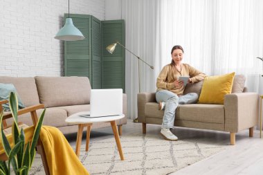 Oturma odasındaki gri koltukta tablet bilgisayar kullanan gülümseyen genç kadın.