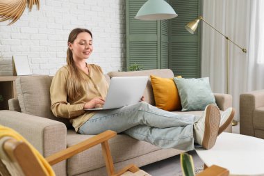 Oturma odasındaki gri koltukta dizüstü bilgisayar kullanan gülümseyen genç kadın.