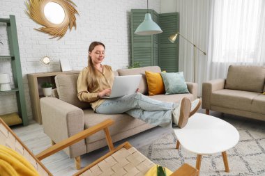 Oturma odasındaki gri koltukta dizüstü bilgisayar kullanan gülümseyen genç kadın.