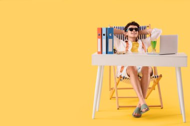 Yaz tatilini bekleyen genç işadamı sarı arka plandaki masada güverte sandalyesinde.