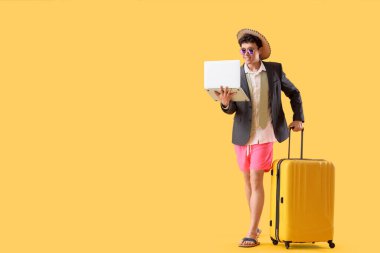 Dizüstü bilgisayarı ve bavulu olan genç işadamı sarı arka planda yaz tatilini bekliyor.