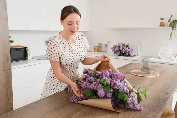Genç bir kadın mutfaktaki masada kağıt ile leylak çiçekleri paketliyor.