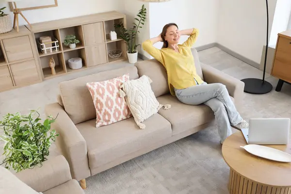 Oturma odasındaki gri kanepede dinlenen gülümseyen genç kadın.