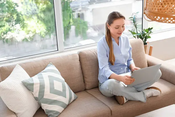 Oturma odasındaki pencerenin yanındaki gri koltukta dizüstü bilgisayar kullanan gülümseyen genç kadın.
