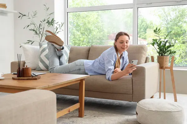 Oturma odasındaki gri koltukta cep telefonu kullanan genç bir kadın.