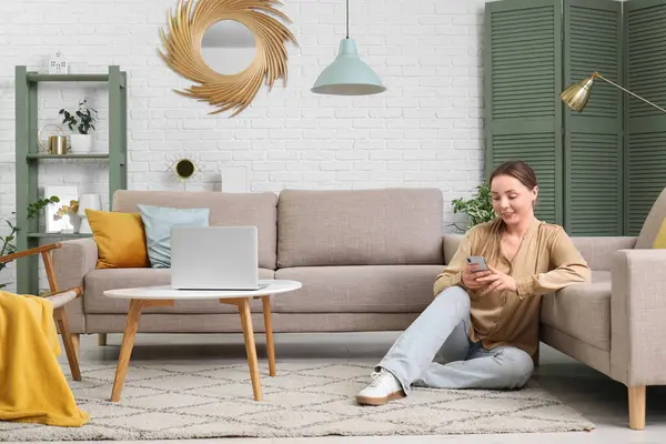 Oturma odasındaki gri kanepenin yanında cep telefonu kullanan gülümseyen genç kadın.