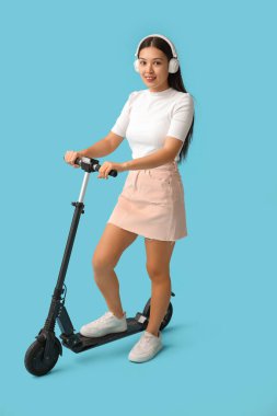 Mavi arka planda modern elektrikli scooterlı kulaklıklı güzel bir Asyalı kadın.