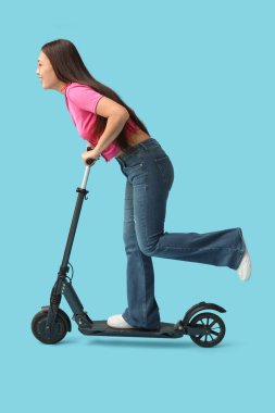 Güzel genç Asyalı kadın mavi arka planda modern elektrikli scooter kullanıyor.