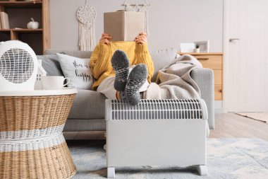 Genç bir kadın oturma odasında kitap okurken modern ısıtıcının üzerinde bacaklarını ısıtıyor.