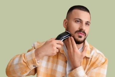 Yeşil arka planda elektrikli traş makinesi olan yakışıklı genç sakallı adam.