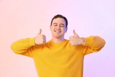 Yakışıklı, genç ve mutlu bir adam. Renkli arka planda baş parmak hareketlerini gösteriyor.
