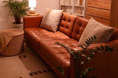 Modern oturma odasının içinde kahverengi kanepe, çekmeceler, ev bitkileri ve akşamları parlayan lamba.