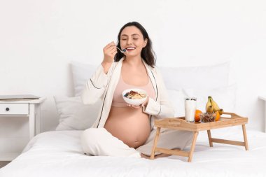 Genç hamile kadın yatak odasında kahvaltı yapıyor.
