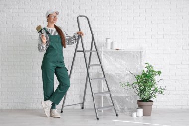 Beyaz tuğla duvarın yanında boya fırçası ve merdiveni olan kadın işçi.