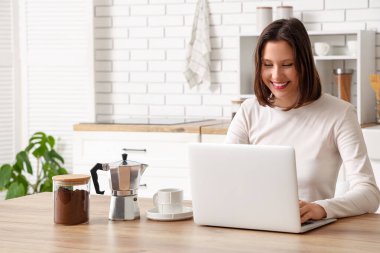 Elinde bir fincan espresso, gayzer kahve makinesi ve mutfaktaki masada oturan dizüstü bilgisayarlı güzel bir kadın.