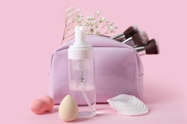 Micellar su, kozmetik çanta, süngerler, pamuk pedler ve pembe arka planda çiçekler.
