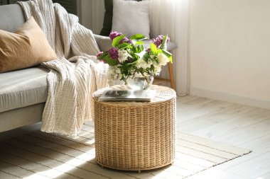 Oturma odasının içinde çiçek açan leylak dalları olan hasır kahve masası.