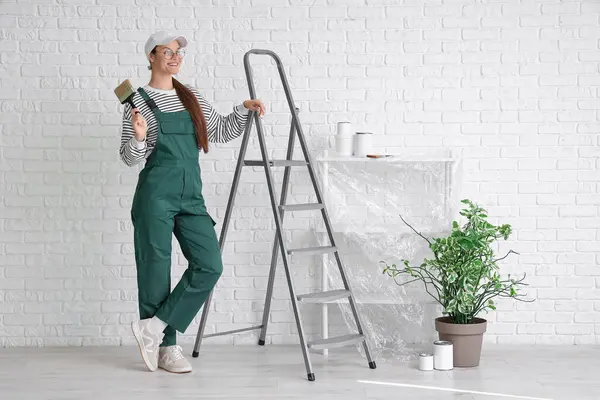Beyaz tuğla duvarın yanında boya fırçası ve merdiveni olan kadın işçi.