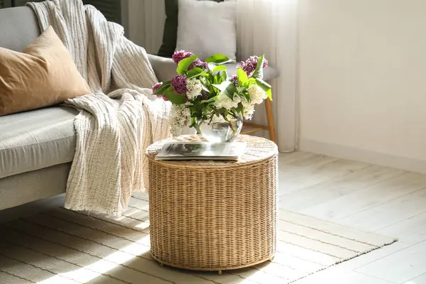 Oturma odasının içinde çiçek açan leylak dalları olan hasır kahve masası.