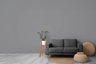 Oturma odasının iç tarafındaki bej duvarın yanında puf, saksı ve gri kanepe.