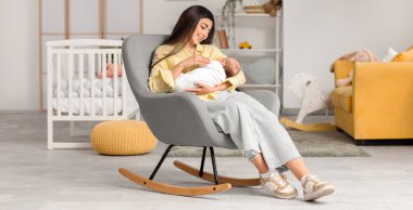 Kadın bebeğini emzirirken evdeki koltukta oturuyor.